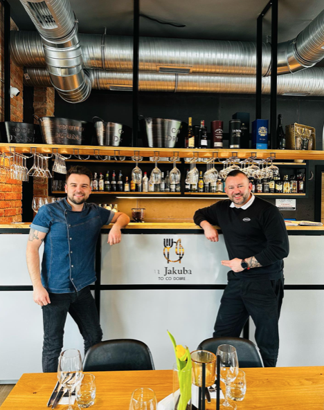 Dwaj uśmiechnięci mężczyźni z obsługi restauracji „u Jakuba” stojący przy barze z logo lokalu w nowoczesnym wnętrzu z ceglaną ścianą.